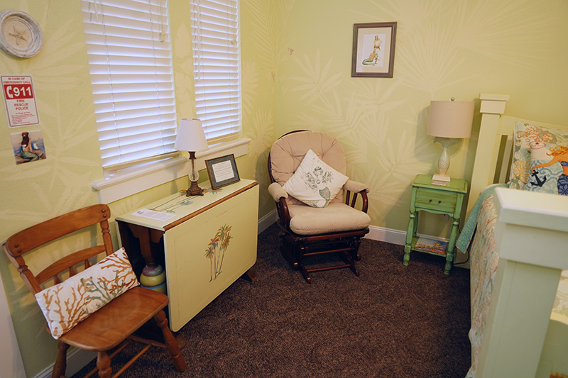 This cozy corner of a bedroom features a rocking chair, a drop-leaf table, and a wooden chair, all set against pale yellow walls with subtle leaf patterns and window blinds providing natural light.