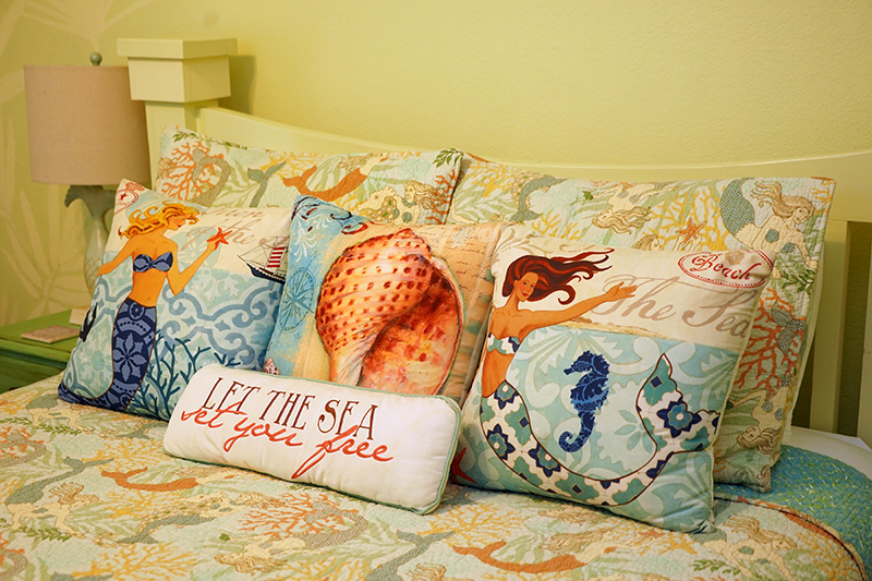 A bed with a vibrant mermaid and sea-themed comforter is adorned with a collection of decorative throw pillows, including a bolster pillow that reads "LET THE SEA SET YOU FREE."