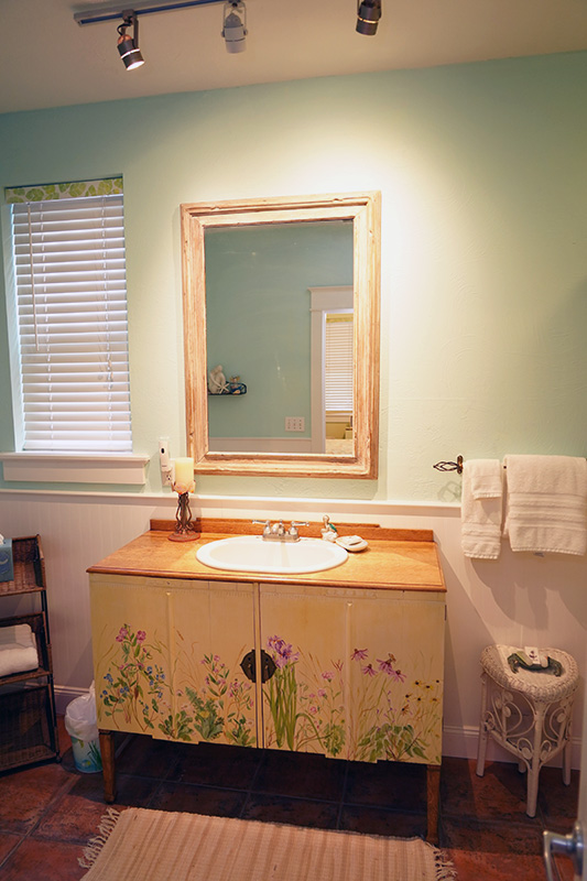 This inviting bathroom features light green walls, a wooden vanity with painted floral details, a large framed mirror, and a window with blinds, all illuminated by track lighting.