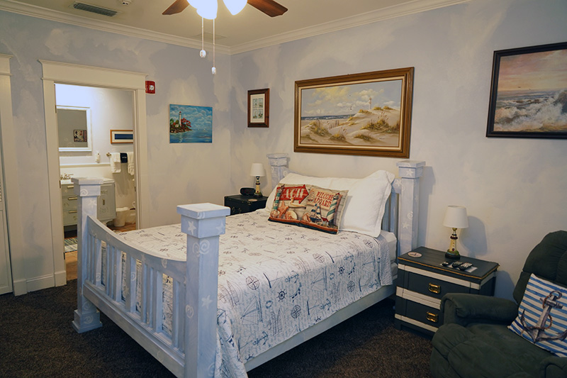 This nautical-themed bedroom features a white wooden bed with a patterned comforter, light blue walls adorned with seascape art, and a view into an adjacent bathroom.