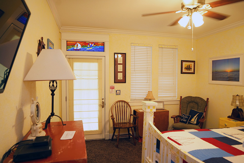 This cheerful bedroom features pale yellow walls, a white bed, a wooden rocking chair, and a door with a colorful stained-glass transom window depicting a house by the sea.