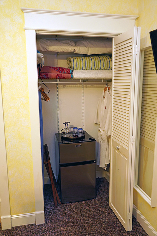 This small, well-organized closet contains a mini-fridge with a hospitality tray, hanging robes, and shelves neatly stacked with extra pillows and blankets.