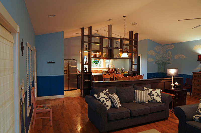 This open-concept living space features light blue walls with darker wainscoting, hardwood floors, a gray sofa, and a wooden room divider that provides a peek into the adjacent kitchen.