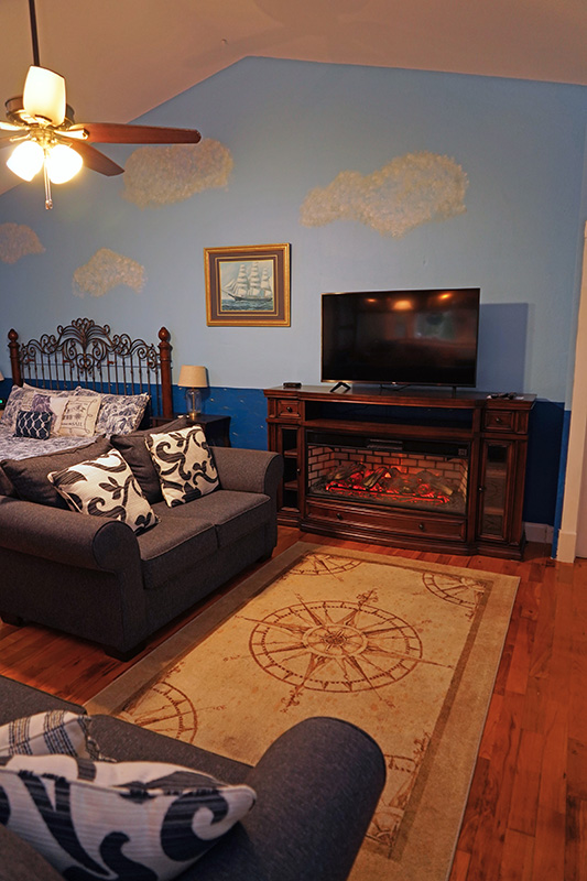 This inviting bedroom features light blue walls with cloud murals, a bed with a dark gray sofa at its foot, a fireplace with a TV above, and a compass rose rug on hardwood floors.
