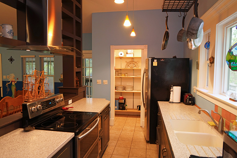 This spacious kitchen features dark wooden cabinetry, light countertops, a black refrigerator, and a pot rack overhead, leading to a well-lit pantry or hallway.