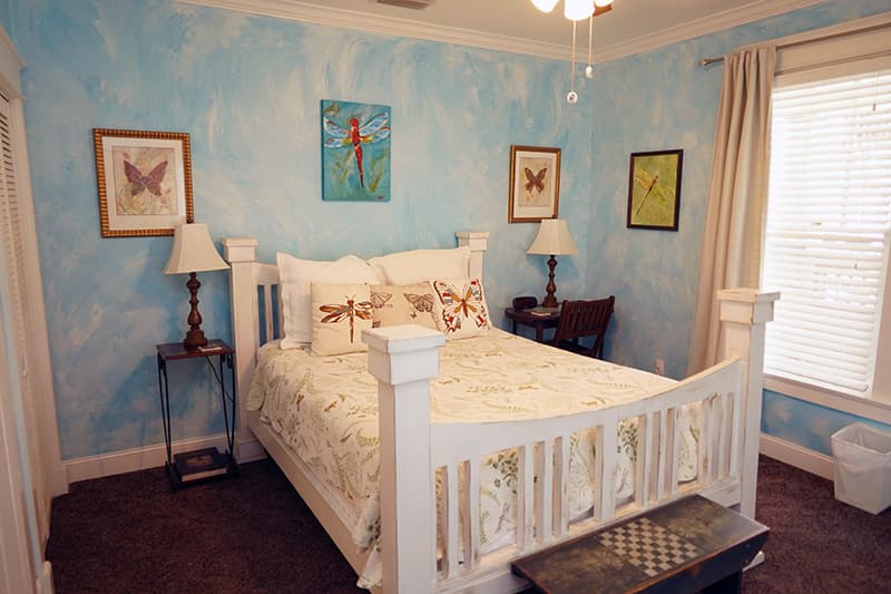 This bright bedroom features a white wooden bed with a floral comforter, light blue textured walls, and butterfly artwork, creating a serene and airy atmosphere.