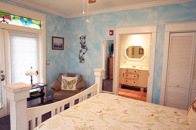 This serene bedroom boasts light blue textured walls, a white wooden bed, a cozy armchair, and a view into an en-suite bathroom with a classic vanity.