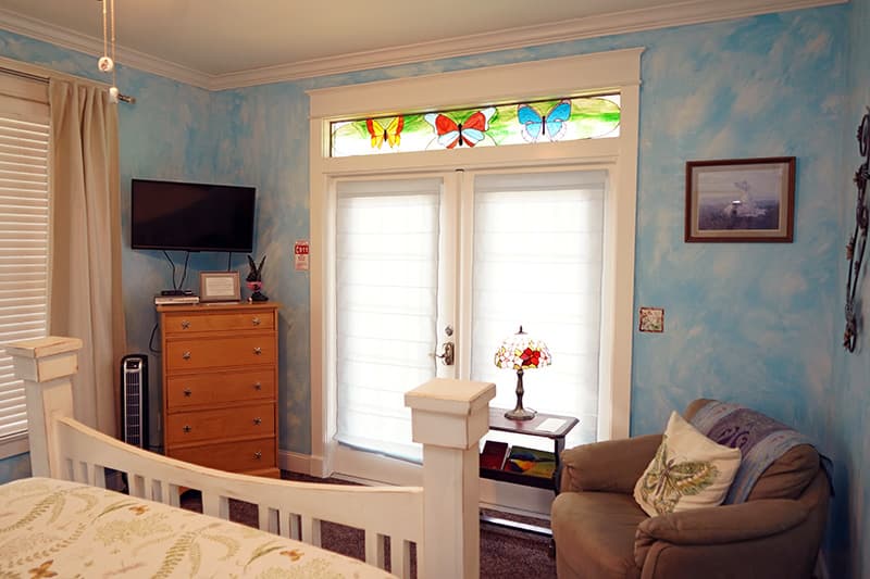 This charming bedroom features blue textured walls, a white bed frame, a cozy armchair, and a doorway topped with vibrant stained-glass butterflies.