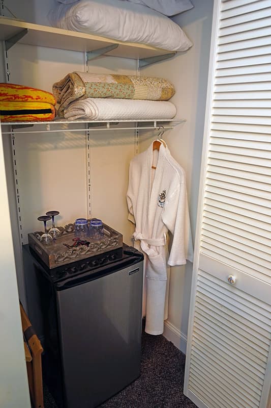 This compact closet offers practical amenities including a mini-fridge with a hospitality tray, a hanging white robe, and neatly folded linens on upper shelves.