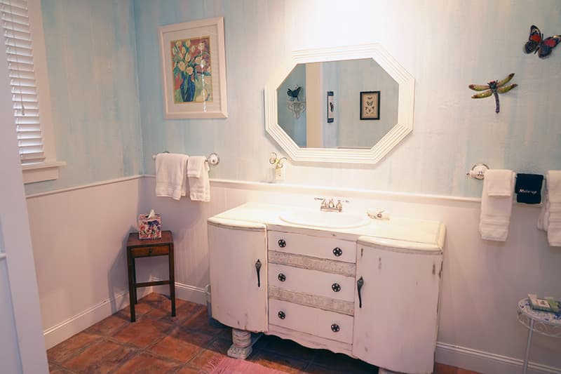 This inviting bathroom features a vintage-style white vanity, a large octagonal mirror, light blue textured walls, and charming dragonfly & butterfly decor.