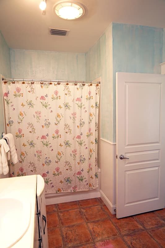 This charming bathroom features light blue textured walls, warm terracotta floor tiles, and a white shower curtain adorned with a colorful floral and butterfly pattern.