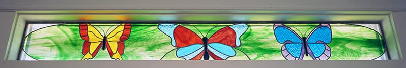 A rectangular stained glass transom window features three vibrant butterflies-yellow, red, and blue-set against a green, leafy background.