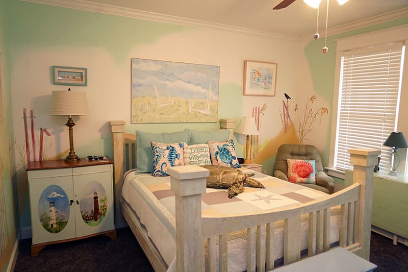 This vibrant, sea-themed bedroom features a white wooden bed, colorful throw pillows, a large stuffed sea turtle, and walls painted with a playful blend of green and white, adorned with whimsical murals.