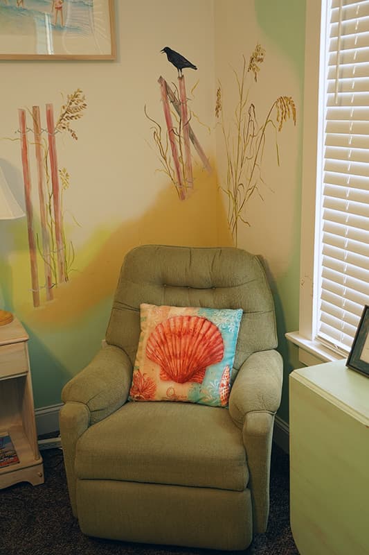 A cozy olive-green armchair with a vibrant seashell throw pillow sits against a wall adorned with a whimsical bird and tall grass mural.