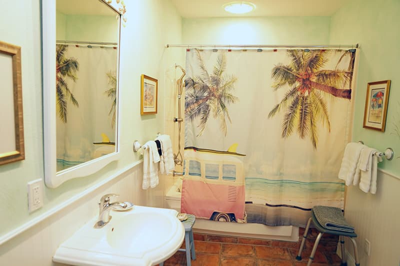This vibrant bathroom features light green walls, a playful shower curtain with palm trees and a retro camper van, a white pedestal sink, and terracotta floor tiles.