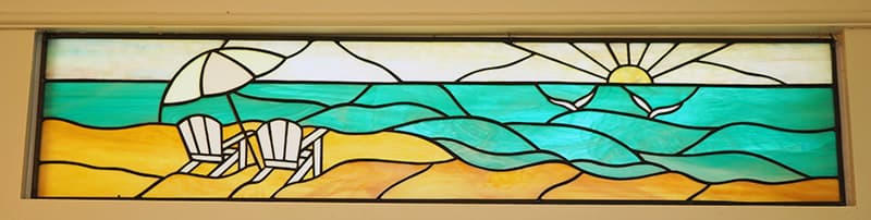 A long, rectangular stained glass panel depicts a serene beach scene with two white Adirondack chairs and an umbrella on the sand, facing turquoise waves and a rising sun in a clear sky.