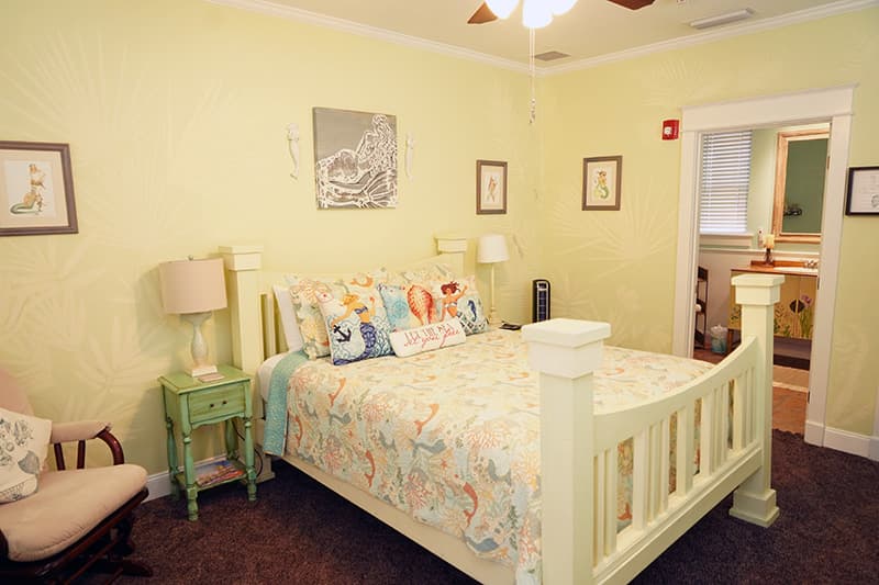This charming bedroom features pale yellow walls, a white wooden bed with colorful bedding, a green nightstand, and an inviting view into an en-suite bathroom.
