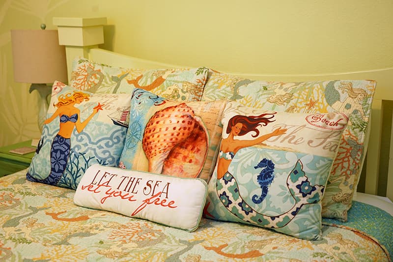 A bed with a vibrant mermaid and sea-themed comforter is adorned with a collection of decorative throw pillows, including a bolster pillow that reads "LET THE SEA SET YOU FREE."