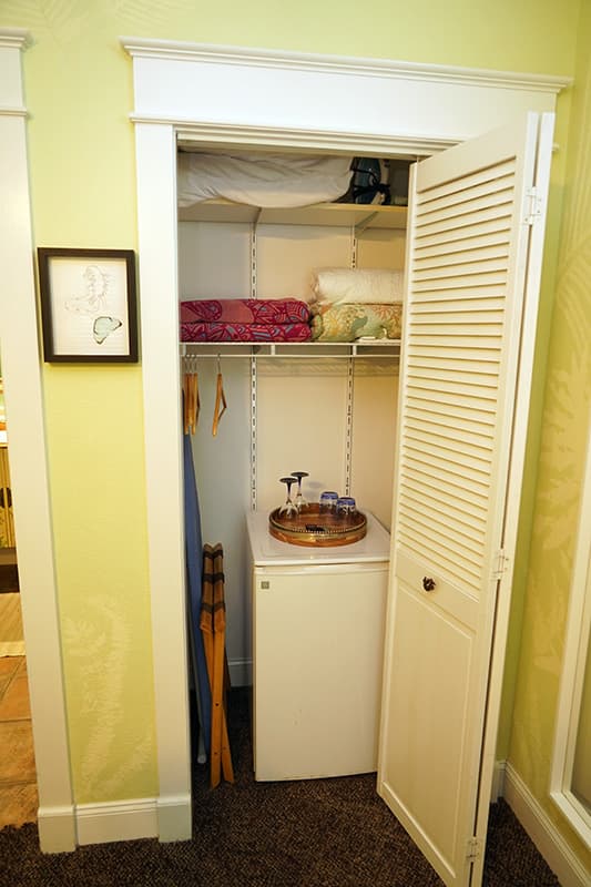 This compact closet, with its louvered bifold doors, houses a mini-fridge topped with a hospitality tray, an ironing board, and shelves stocked with extra linens and towels.