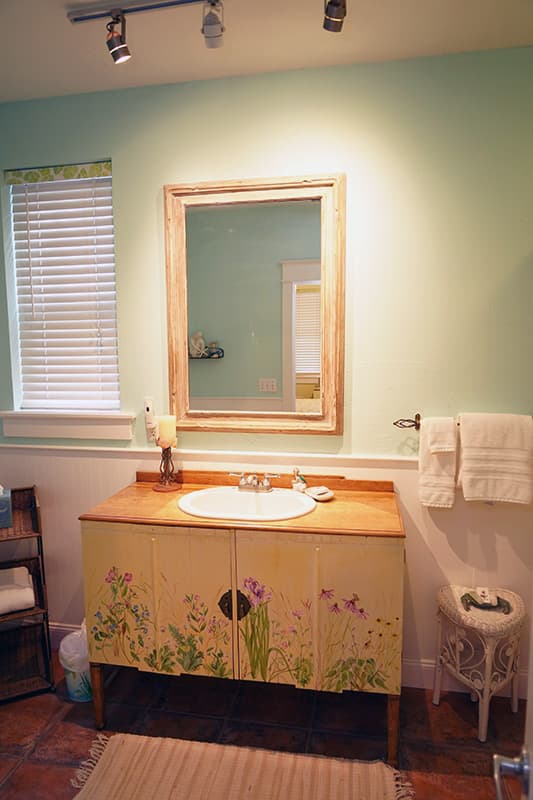 This inviting bathroom features light green walls, a wooden vanity with painted floral details, a large framed mirror, and a window with blinds, all illuminated by track lighting.
