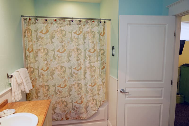 This light-filled bathroom features a shower with a whimsical patterned curtain, pale green and white walls, and a glimpse into an adjacent room through an open white door.