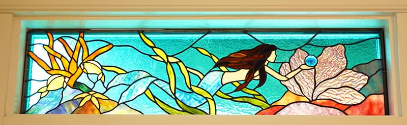 A horizontal stained glass panel depicts an underwater scene with a mermaid, vibrant aquatic plants, and large flowers, all set against a shimmering turquoise background.