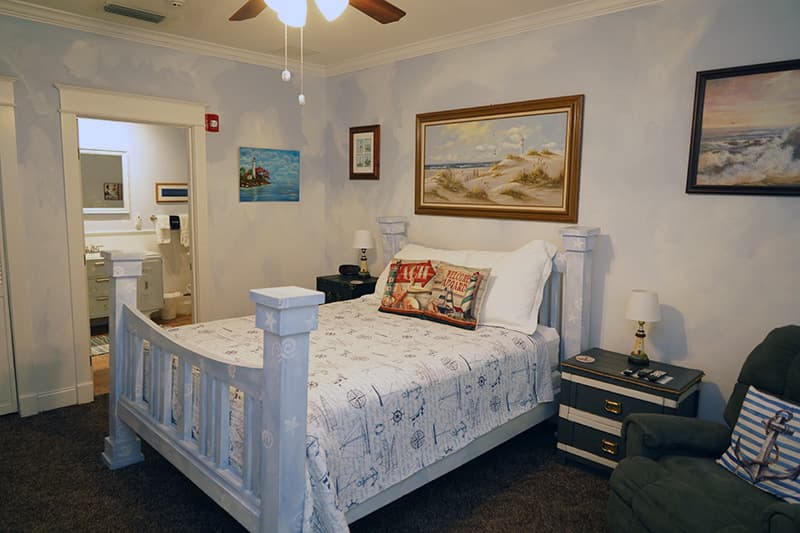 This nautical-themed bedroom features a white wooden bed with a patterned comforter, light blue walls adorned with seascape art, and a view into an adjacent bathroom.