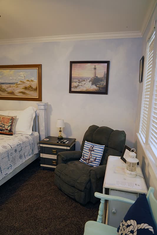 This corner of a nautical-themed bedroom features an olive-green recliner with an anchor pillow, a white nightstand, and framed lighthouse artwork on the light blue wall.