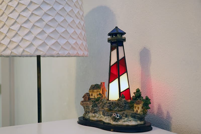 A detailed lighthouse lamp, featuring red and white stripes, stands on a white surface next to a lamp with a textured white shade, casting a warm glow.