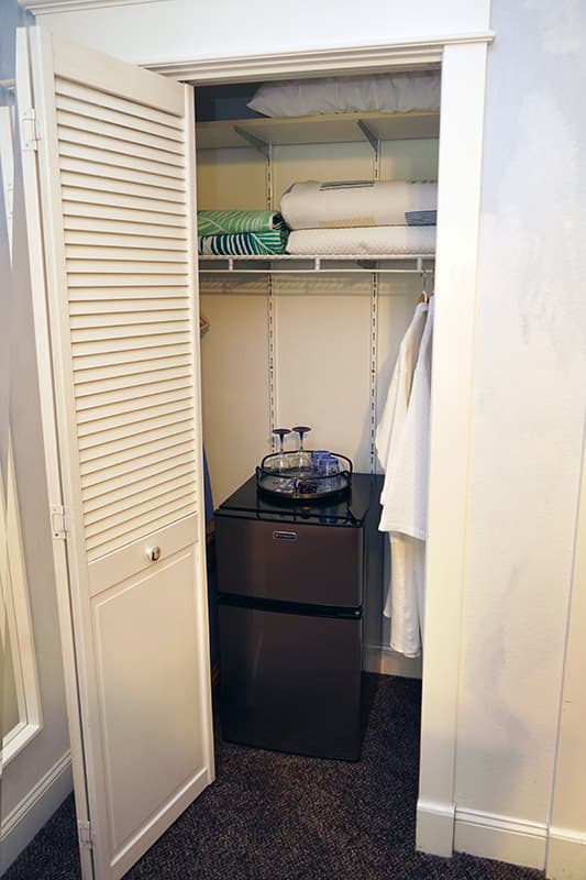 This well-equipped closet features louvered bifold doors, a compact refrigerator with a hospitality tray, and shelves neatly stocked with linens and a hanging robe.