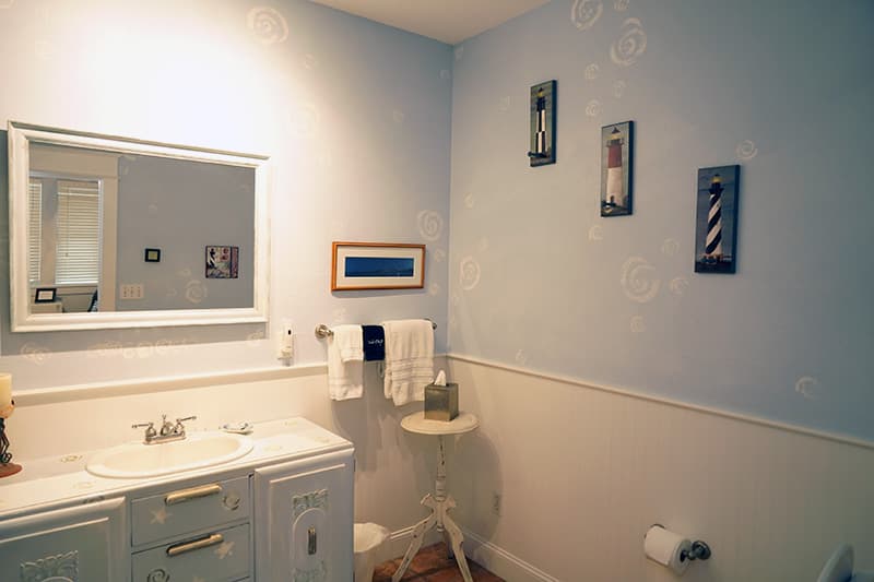 This charming nautical-themed bathroom features light blue walls with subtle white patterns, a light blue vanity, a framed mirror, and decorative wall art including three lighthouse motifs.