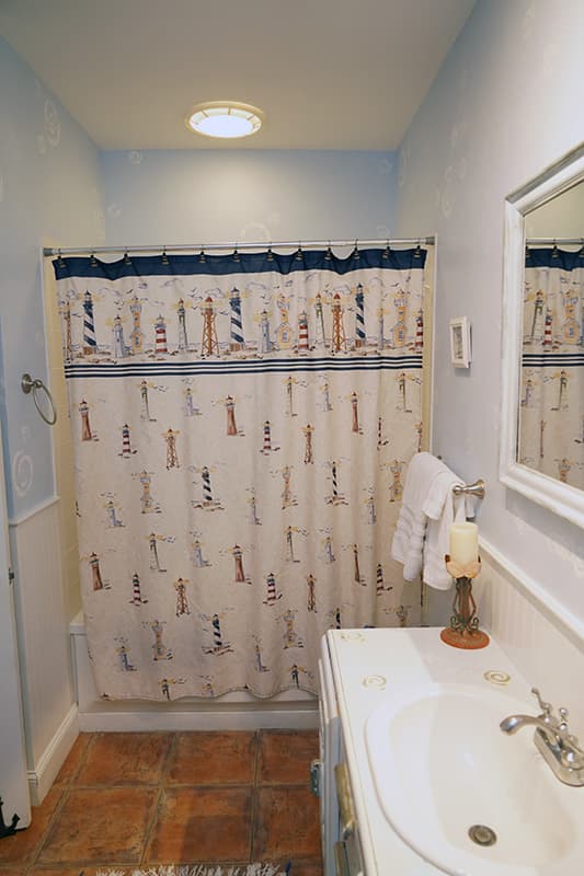 This cozy bathroom features a nautical-themed shower curtain with lighthouses, light blue walls, a white vanity with a sink, and terracotta-colored floor tiles.