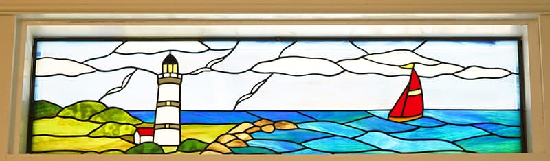 A stained glass panel depicts a coastal scene with a white and red lighthouse on a green and yellow shore, facing a red-sailed sailboat on a blue, choppy sea under a cloudy sky.