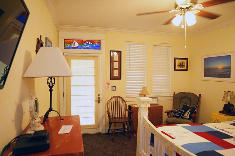 This cheerful bedroom features pale yellow walls, a white bed, a wooden rocking chair, and a door with a colorful stained-glass transom window depicting a house by the sea.