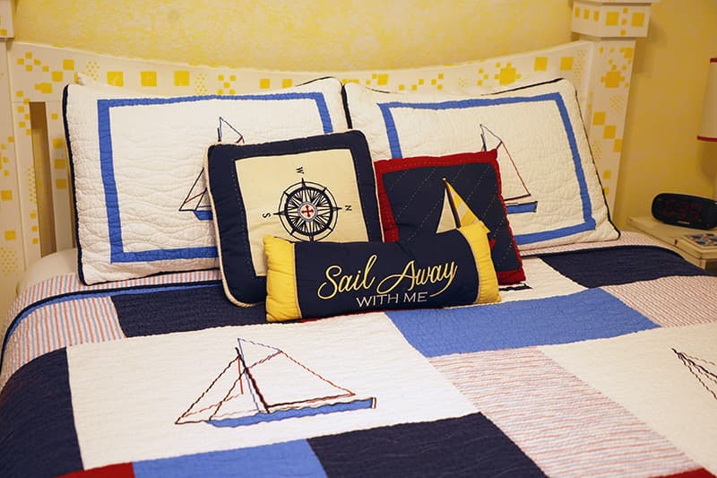 A cozy bed features nautical-themed bedding with a patchwork comforter, two standard pillows with sailboat designs, and a collection of decorative throw pillows, including one that reads "Sail Away With Me."