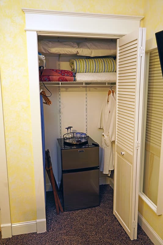 This small, well-organized closet contains a mini-fridge with a hospitality tray, hanging robes, and shelves neatly stacked with extra pillows and blankets.