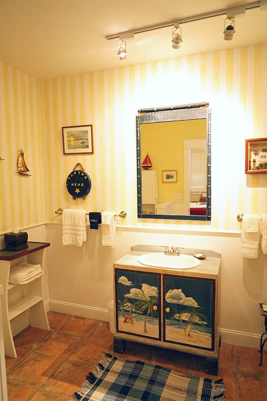 This bright bathroom features vertical yellow and white striped wallpaper, a vanity with beach-themed painted cabinet doors, a large mirror, and terracotta floor tiles with a blue plaid rug.