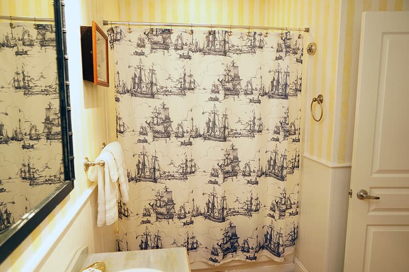 This charming bathroom features yellow and white striped walls, a white shower curtain with a blue sailboat pattern, and a glimpse of a vanity mirror on the left.
