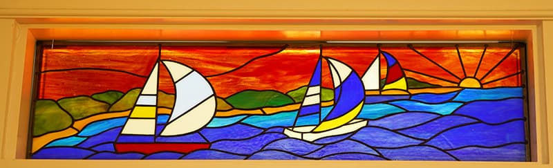 A vibrant stained glass panel depicts three sailboats with colorful sails on a blue sea, against a backdrop of green land and a fiery orange and red sunset with a radiating sun.