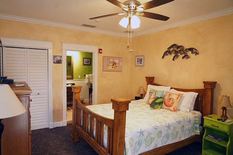 This inviting bedroom features warm, textured yellow walls, a wooden bed with a cheerful patterned comforter, and a clear view into a bright green en-suite bathroom.