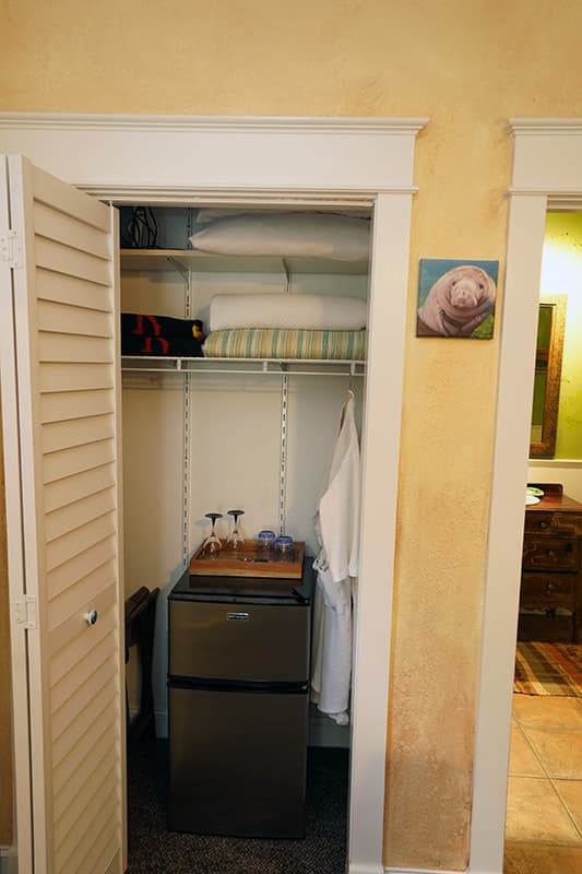 This open closet reveals a mini-fridge with a hospitality tray, a hanging white robe, and shelves stocked with extra linens, all neatly arranged for guest convenience.