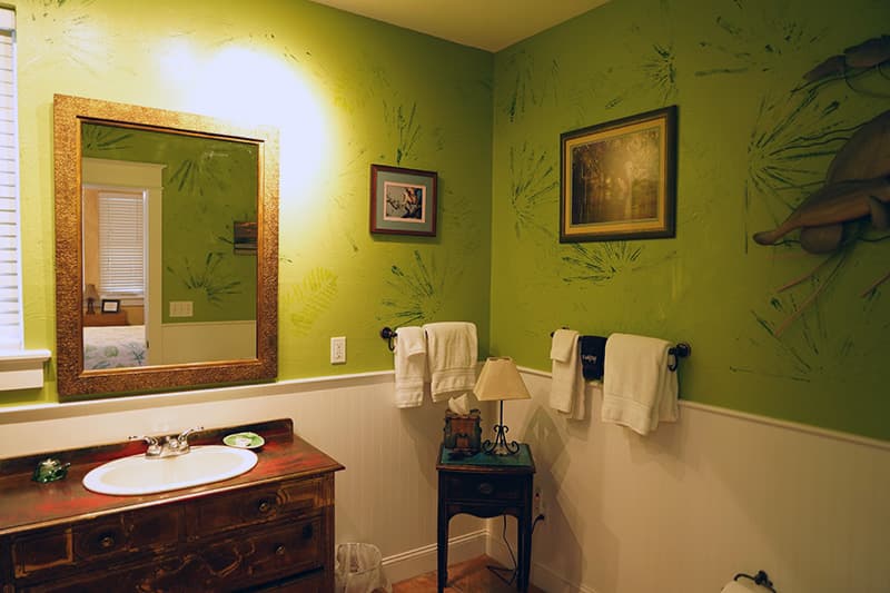 This vibrant bathroom features bright green walls with subtle plant-like patterns, a distressed wooden vanity with a white sink, and a large framed mirror reflecting a portion of the room.