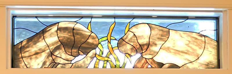 A rectangular stained glass panel depicts two brown manatees, one on each side, with swirling yellow and green shapes in the center, all against a blue background.
