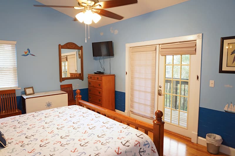 This nautical-themed bedroom features a wooden bed with anchor bedding, light blue walls with a darker blue wainscoting, and French doors leading to an outdoor area.