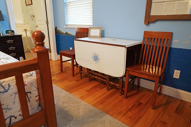 This inviting corner of a bedroom features a drop-leaf table with a compass rose design, flanked by two wooden chairs, all on hardwood floors against light blue walls.