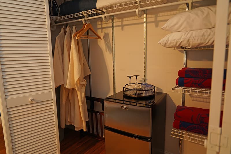 This well-organized closet features a mini-fridge with a hospitality tray, hanging robes, and shelves stocked with neatly folded towels and pillows, offering guest convenience.