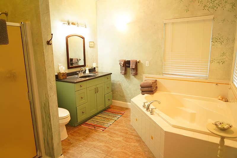 This tranquil bathroom features pale green walls with subtle botanical patterns, a green vanity, a spacious corner whirlpool tub, and a shower stall, all on terracotta-hued floor tiles.