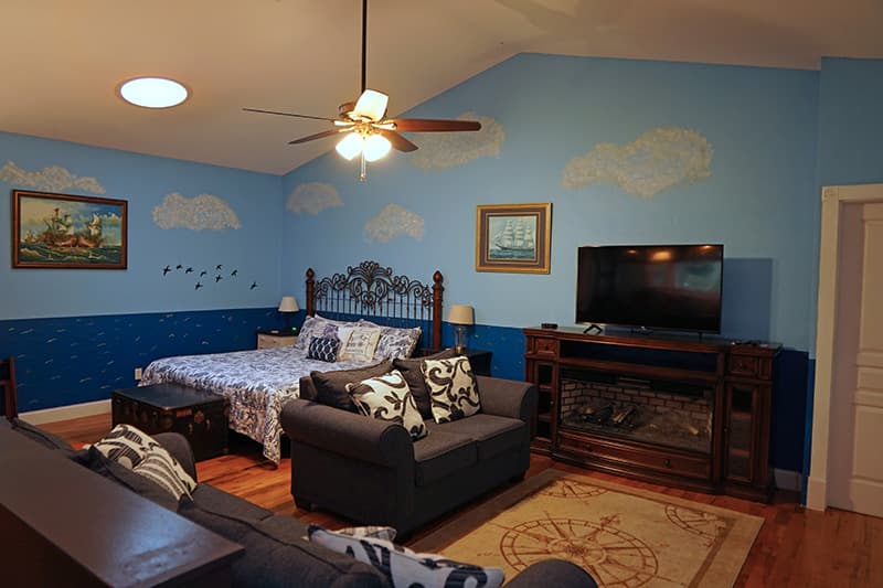 This nautically themed bedroom features light blue walls with cloud murals, a large bed, two sofas, a fireplace, and a television on hardwood floors.