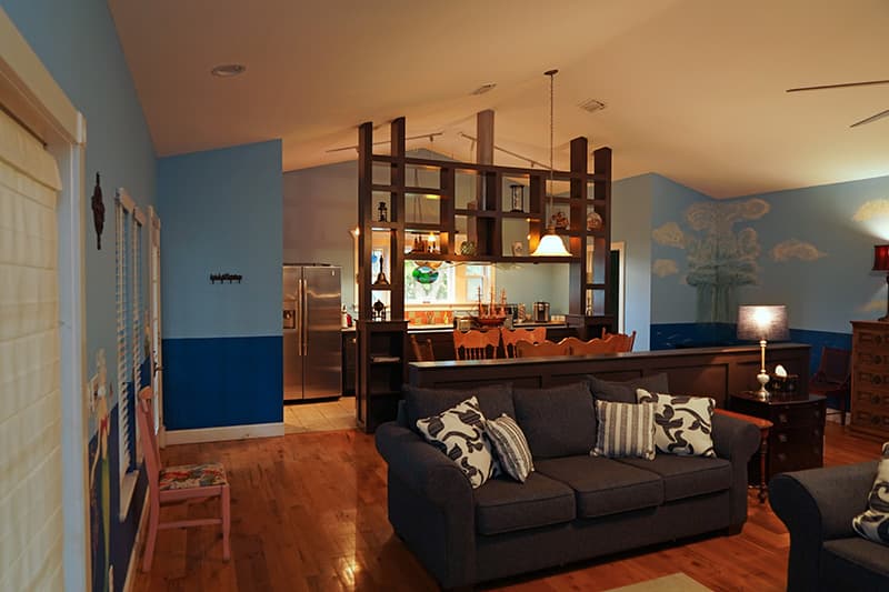 This open-concept living space features light blue walls with darker wainscoting, hardwood floors, a gray sofa, and a wooden room divider that provides a peek into the adjacent kitchen.
