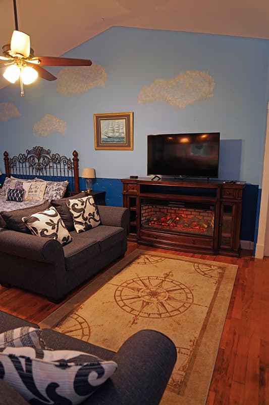 This inviting bedroom features light blue walls with cloud murals, a bed with a dark gray sofa at its foot, a fireplace with a TV above, and a compass rose rug on hardwood floors.